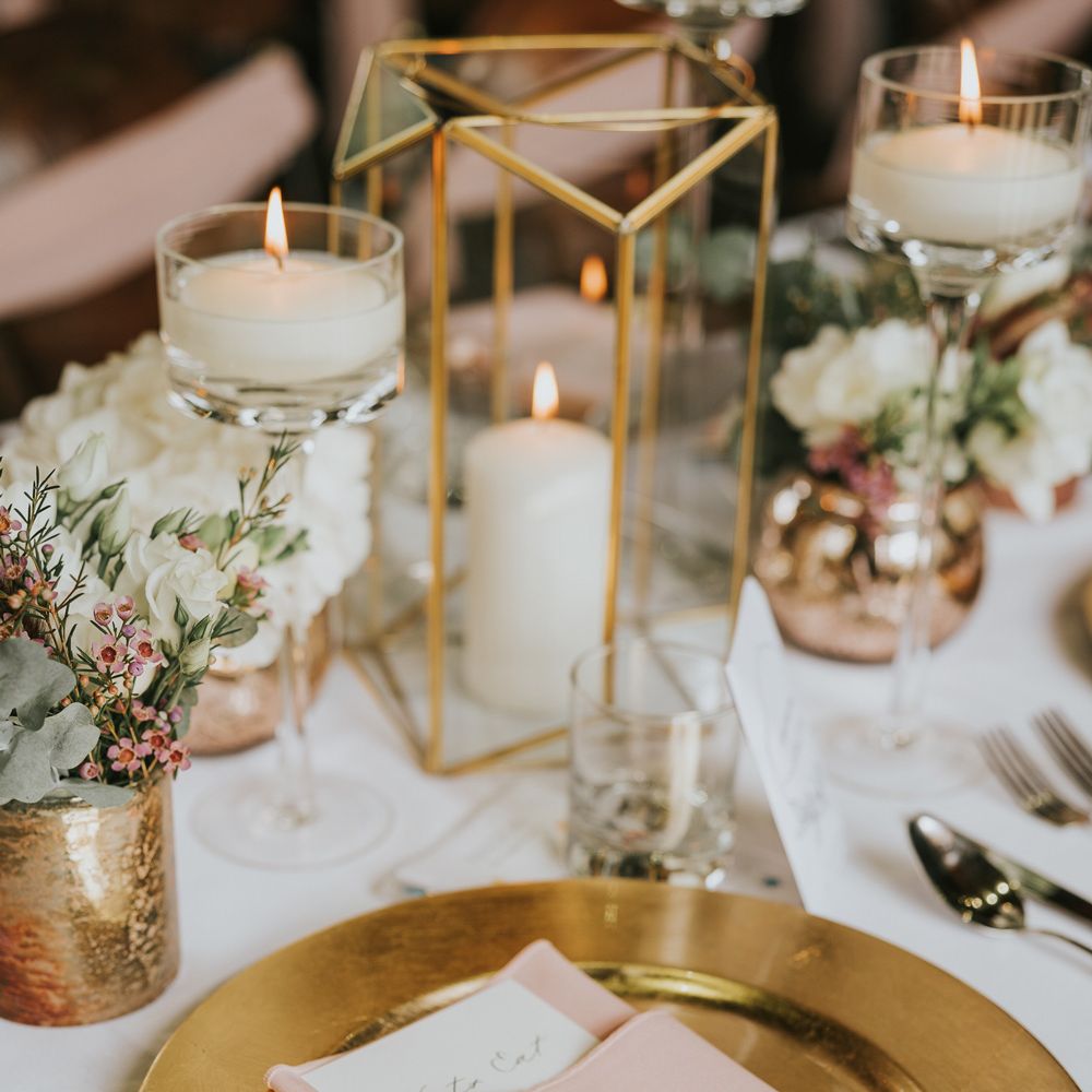 Gold plate with pink napkin and frosted acrylic table place name next to candles, foliage and cutlery for wedding breakfast at Tythe Barn wedding with barn wedding flowers