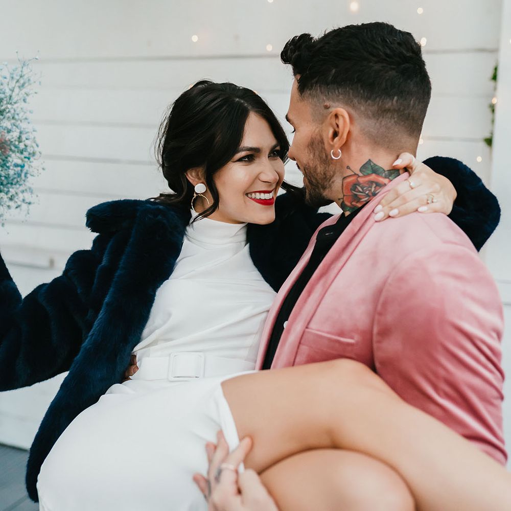 Groom in a pink velvet jacket picking up his bride in a halterneck wedding dress with front split wearing a navy faux fur coverup and red lipstick 