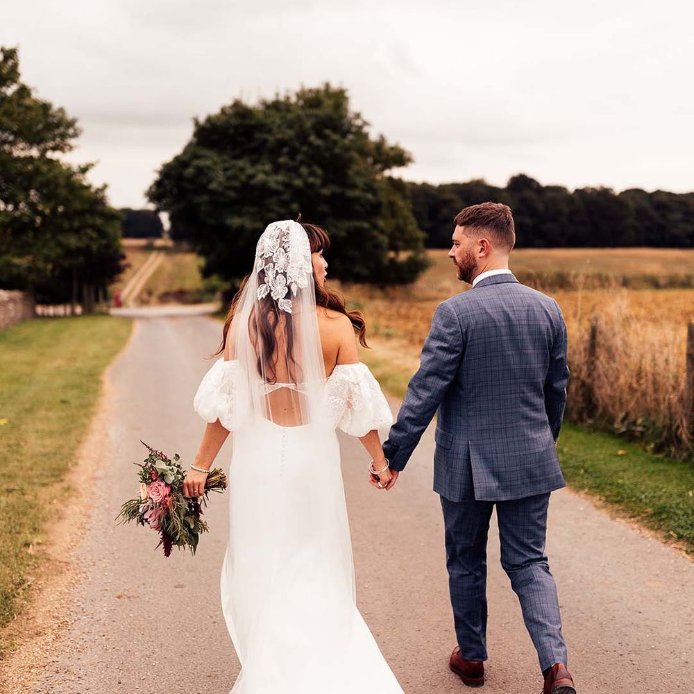 Bride & groom walk hand in hand through the countryside at the Stone Barn Cotswolds on their wedding day