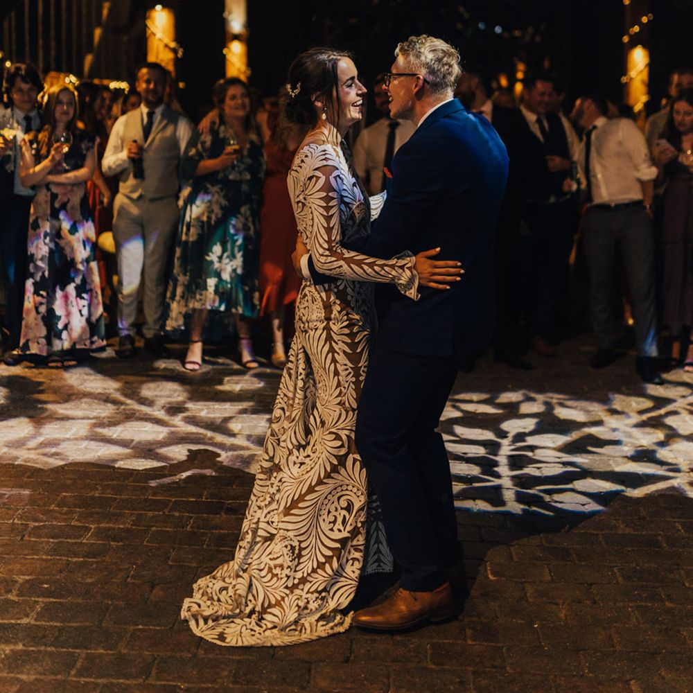 Bride dances in her lace wedding dress with Groom in his three piece dark suit while wedding guests smile under the fairly lights in Barn setting