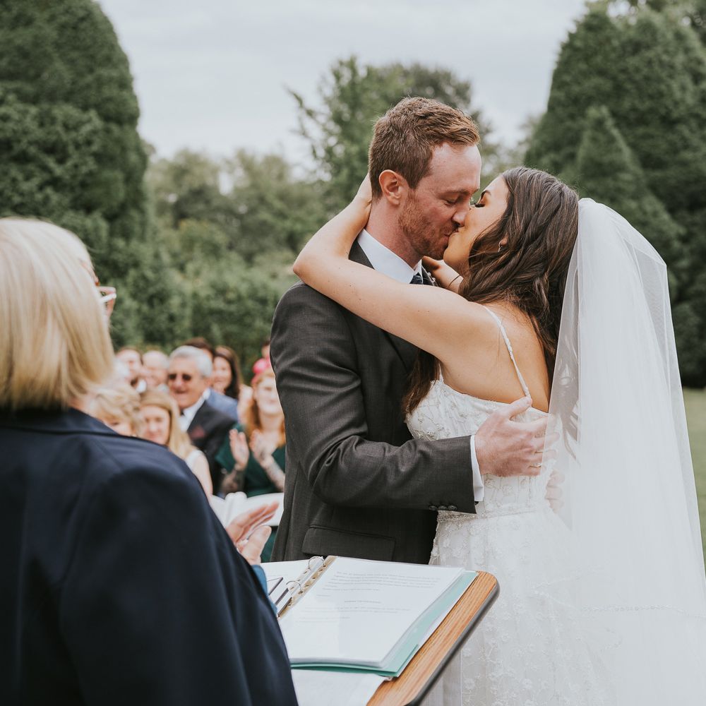 Bride in white cami strap Pronovias wedding dress and veil kisses groom min grey three piece suit during outdoor wedding ceremony at Notley Abbey
