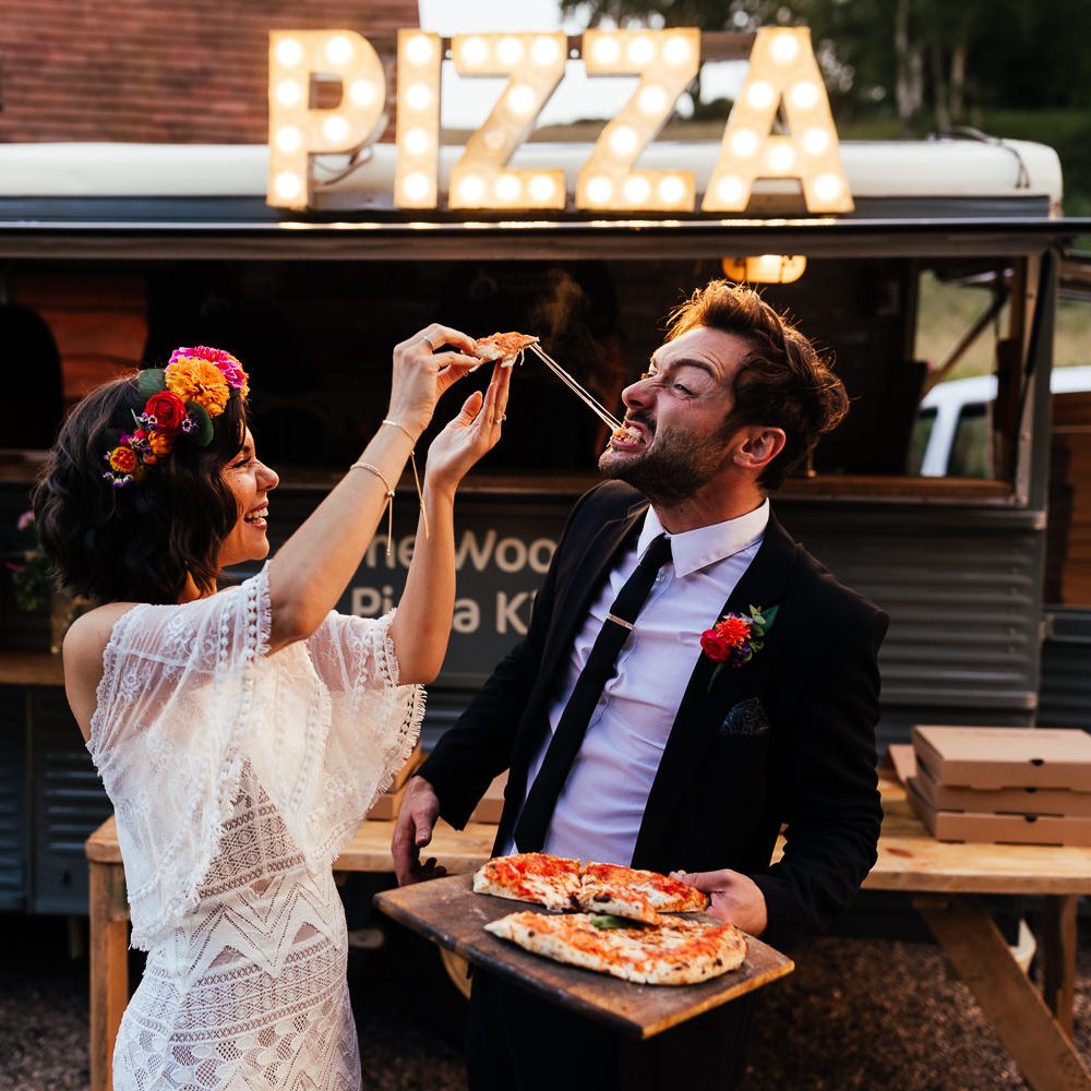 Bride in strapless Grace Loves Lace wedding dress feeds groom in black suit a slice of Pizza by pizza van at rustic barn wedding reception