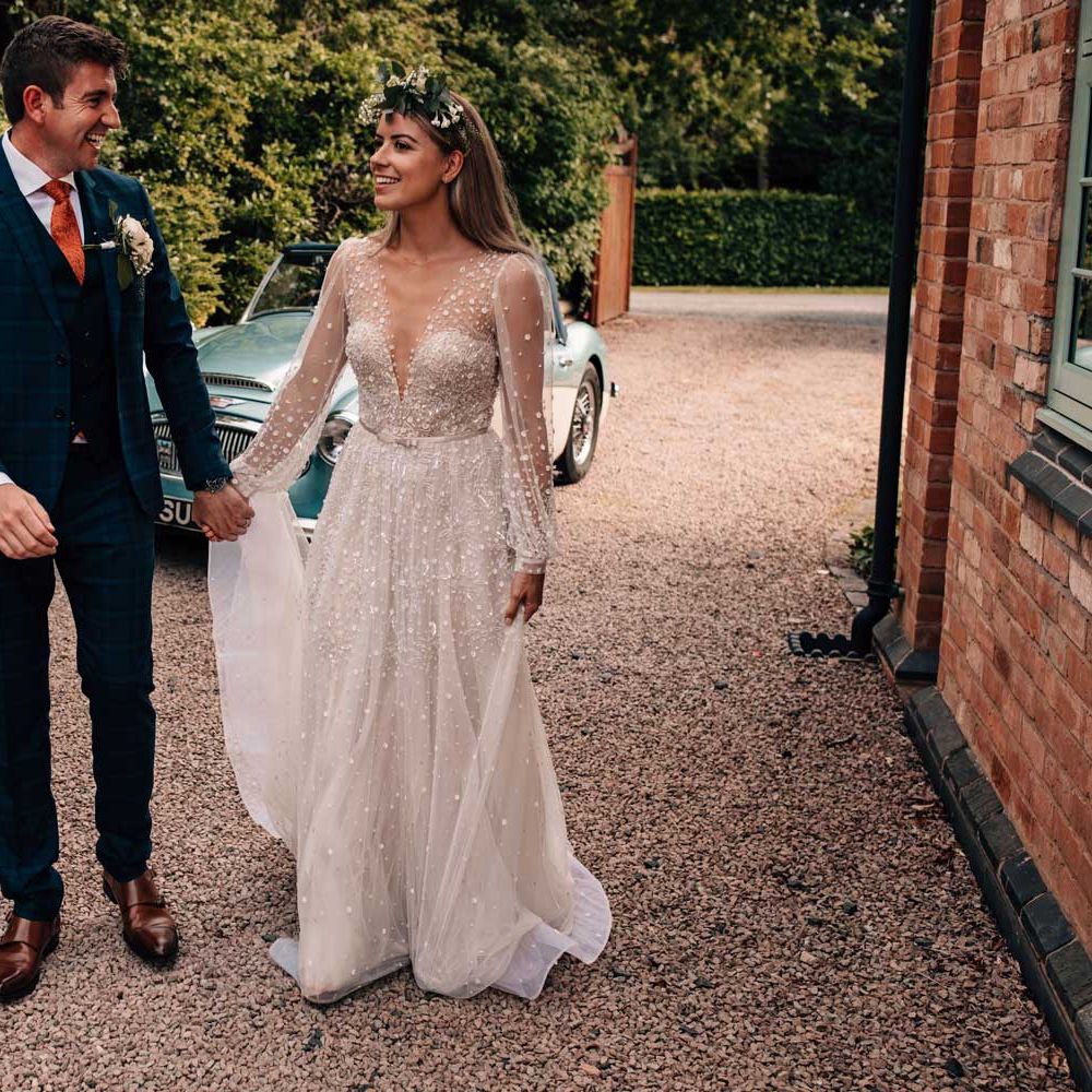 Bride in v neck long sleeve Julie Vino wedding dress and flower crown walks across drive with groom in blue checked three piece suit and floral button hole