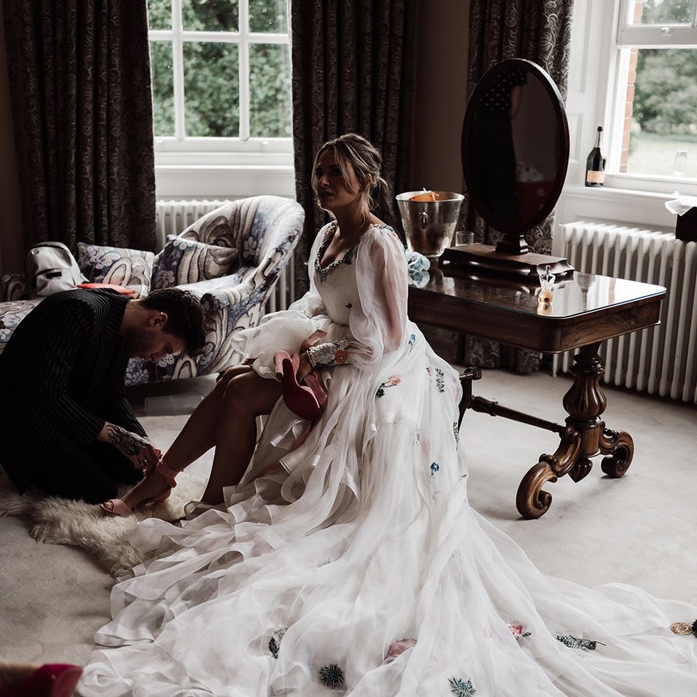 Bride with ponytail wearing long sleeve Victoria Sanders wedding dress with train and floral embroidery sits on stool whilst having her shoe tied