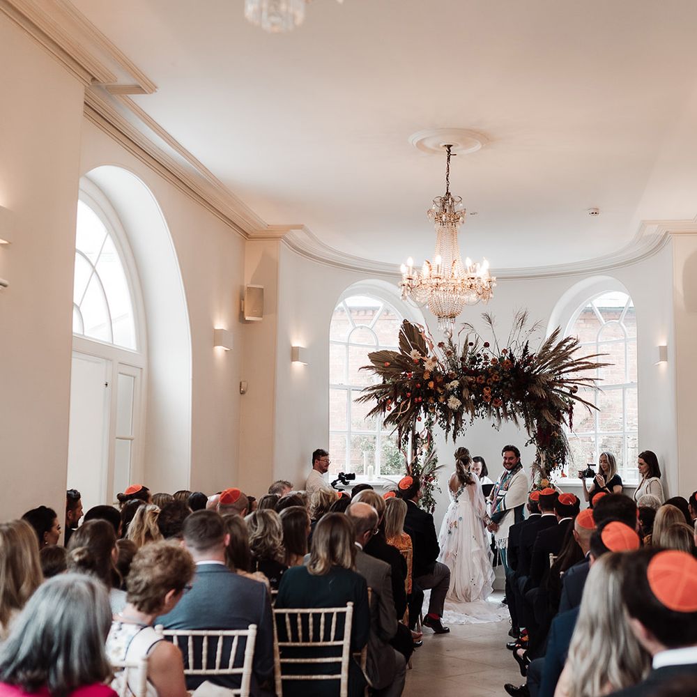 Bride in long mesh sleeve wedding dress with floral embroidery stands with groom in kippah and green suit under arch with dried flower cloud for interfaith wedding ceremony at Iscoyd Park