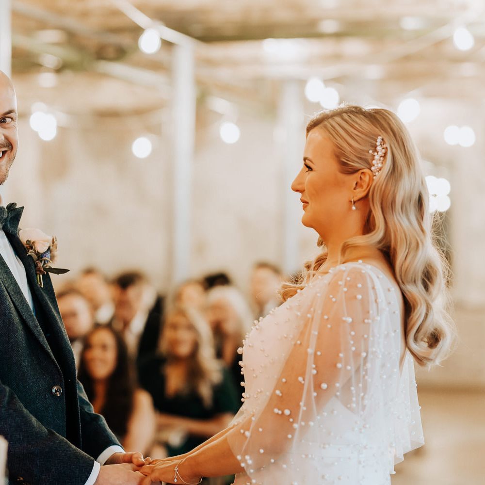 Bride in pearl wedding dress with pearl drop earrings and pearl hair slider stands with groom in navy tweed suit with bow tie and buttonhole during wedding ceremony