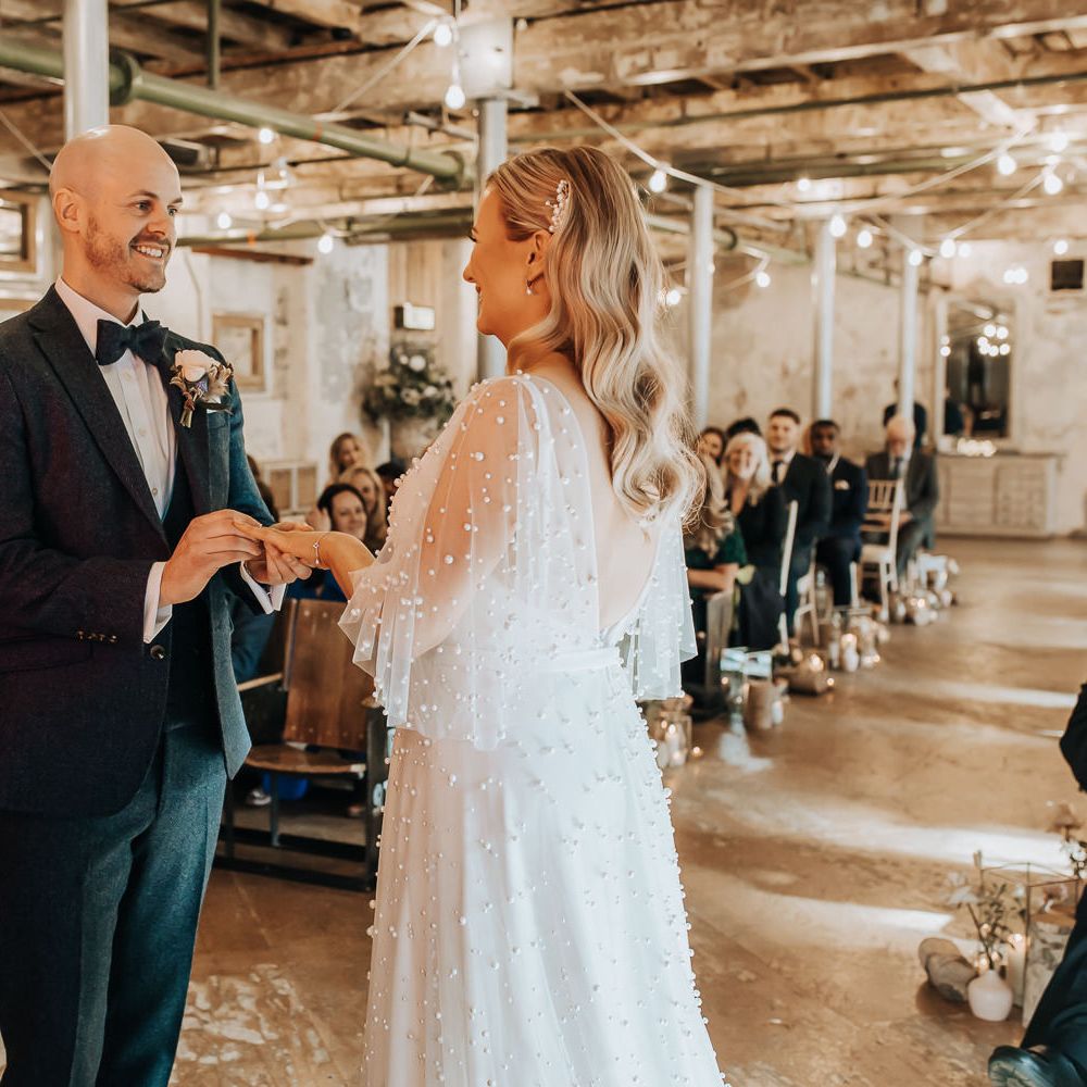 Groom in navy suit and bow tie puts ring on finger of bride in low back pearl wedding dress with sleeves and pearl hair clip during wedding ceremony