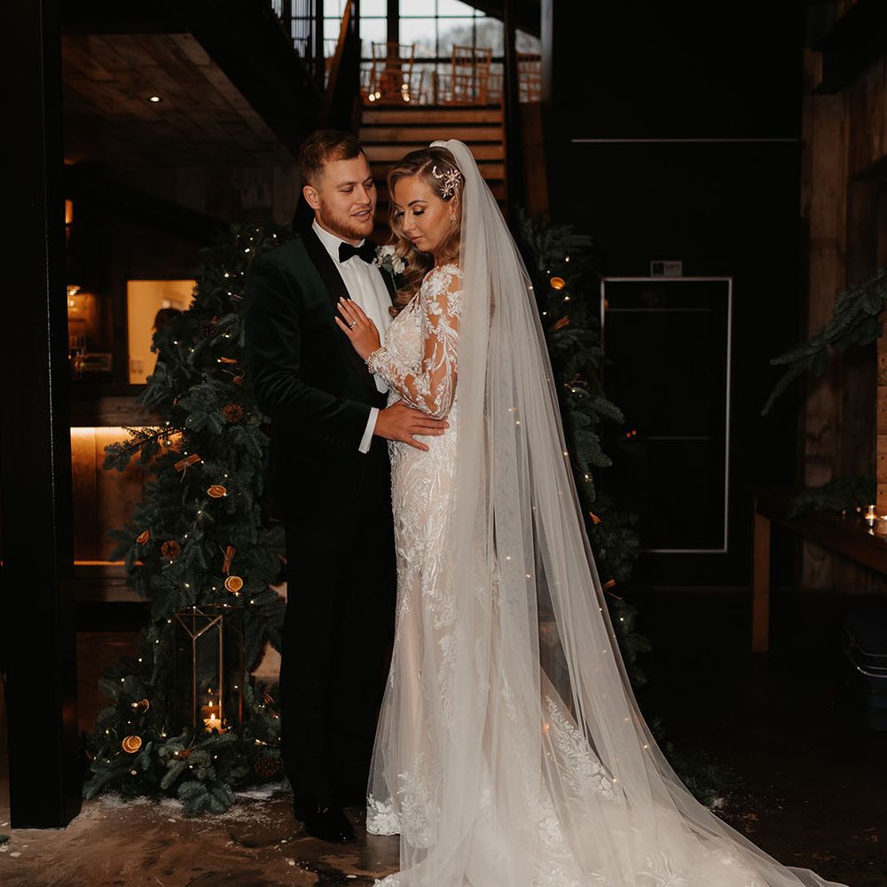 Bride in lace wedding gown and groom in black tie stand in front of dried orange decorated stairway on wedding day