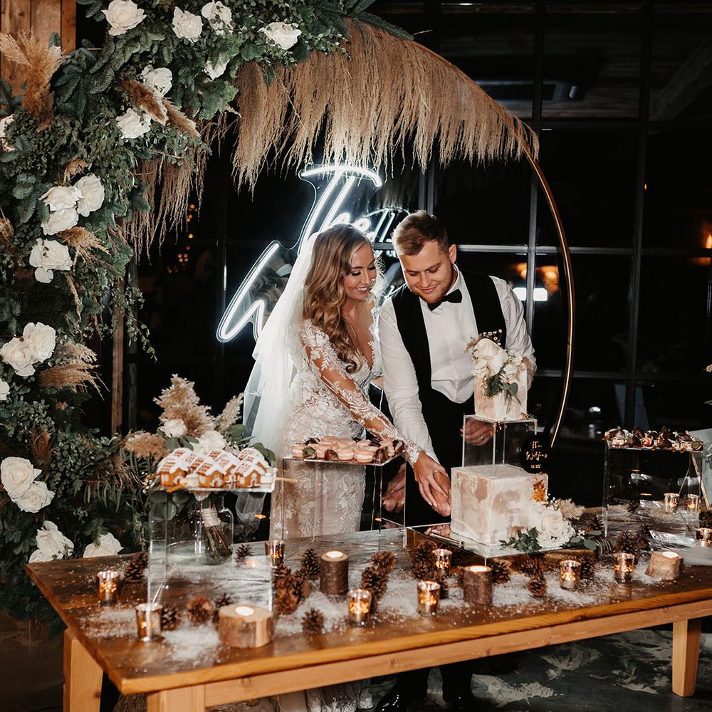 Bride and groom in front of neon sign cut their modern wedding cake next to pink macarons and pinecone and tree trunk candle holders 
