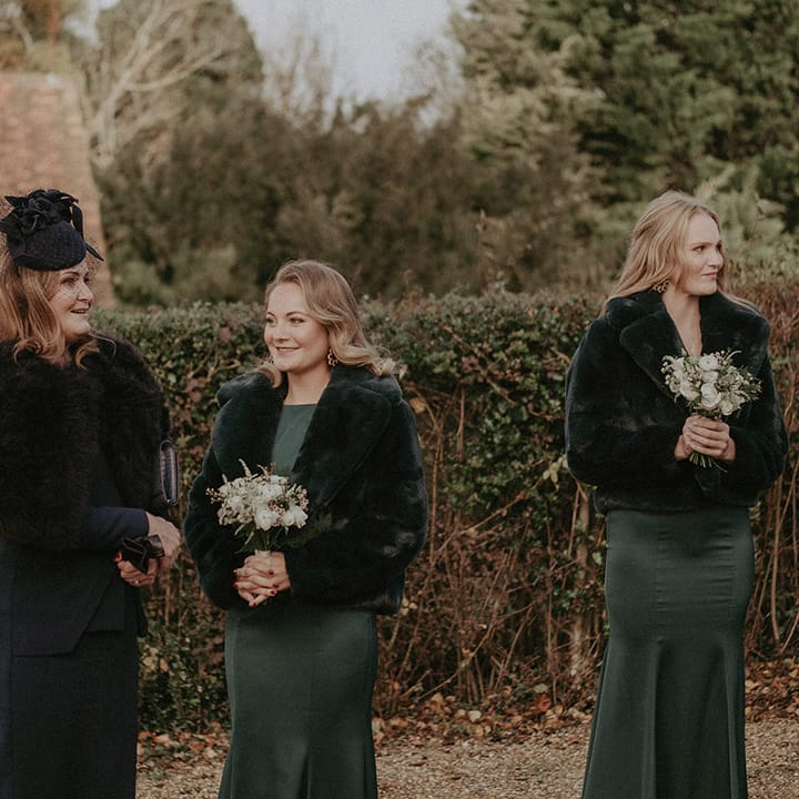 Bridesmaids in dark and bottle green bridesmaids dresses with mixed floral bouquets, black cropped fur jackets and black hats
