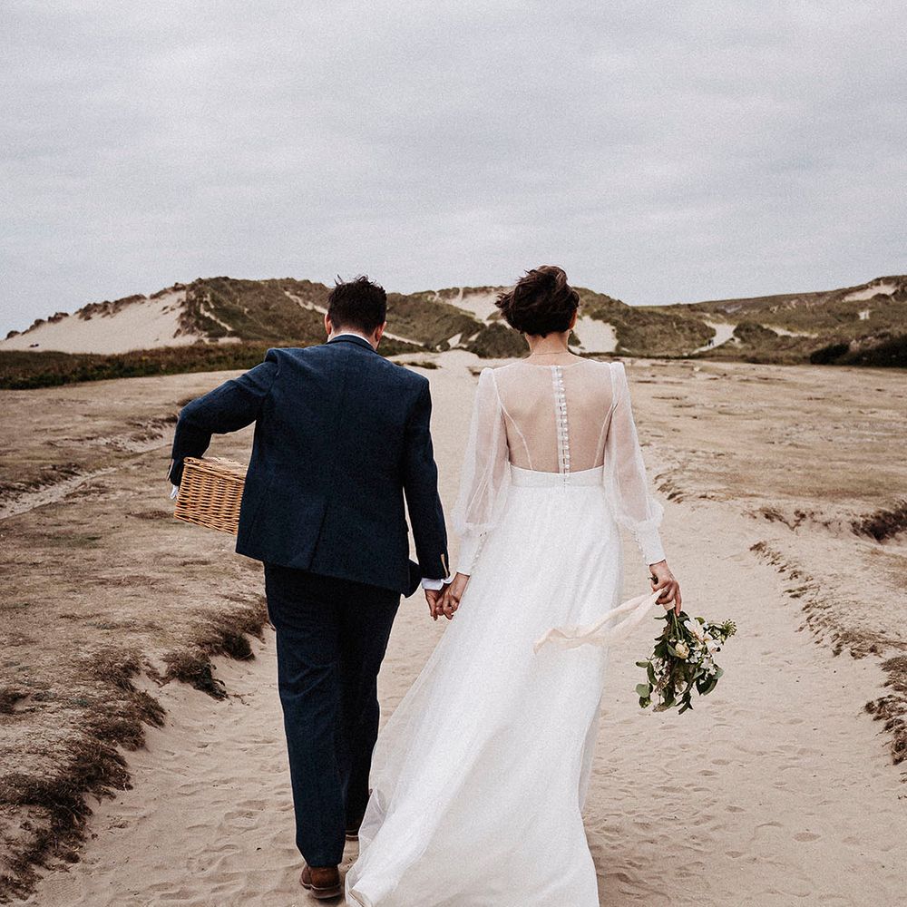 Bride and groom walk along the beach at Cornwall during their wedding day with the groom carrying a wicker picnic basket and the bride holding a wedding bouquet 