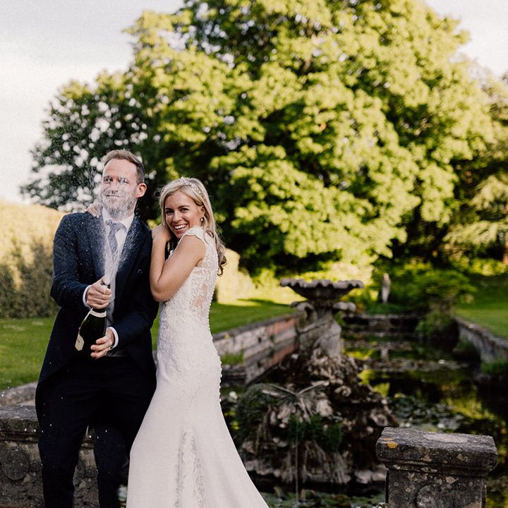 Groom in morning suit pops a bottle of champagne with bride leaning against him in lace wedding dress and pearl headpiece 