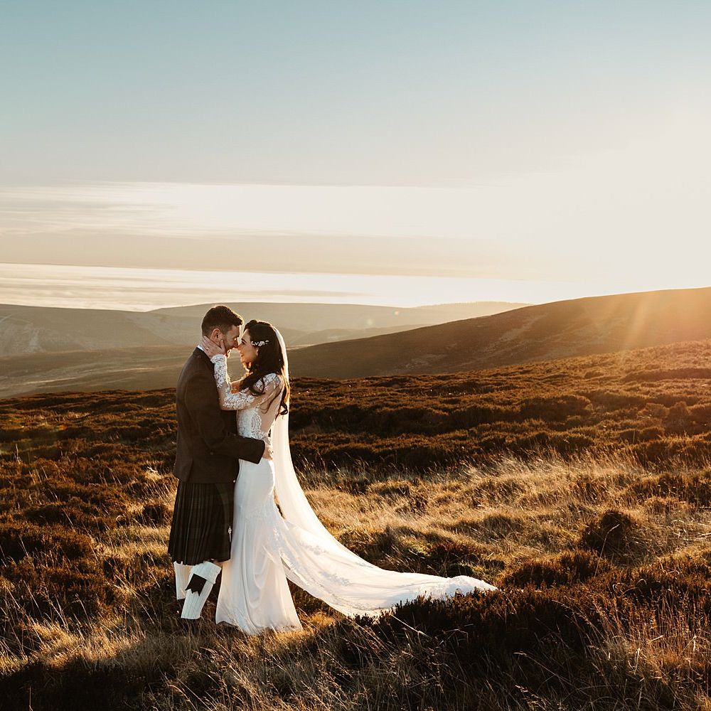 Bride & groom stand on hillside in front of beautiful Scottish highlands whilst the sun sets 