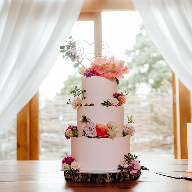 Three tier pink frosted wedding cake complete with floral decor and silver personalised cake topper on wooden cake stand