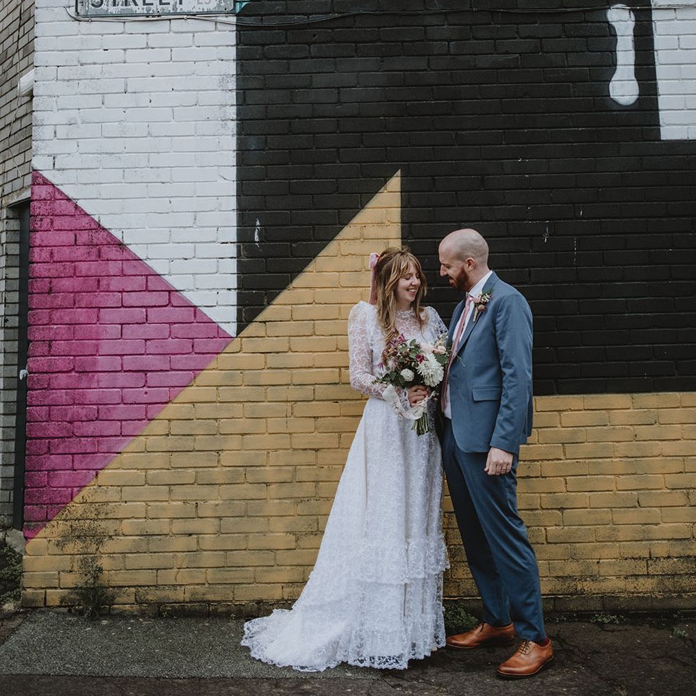 Bride wearing lace wedding dress stands with her groom in front of geometric painted brightly coloured brick wall in Liverpool 