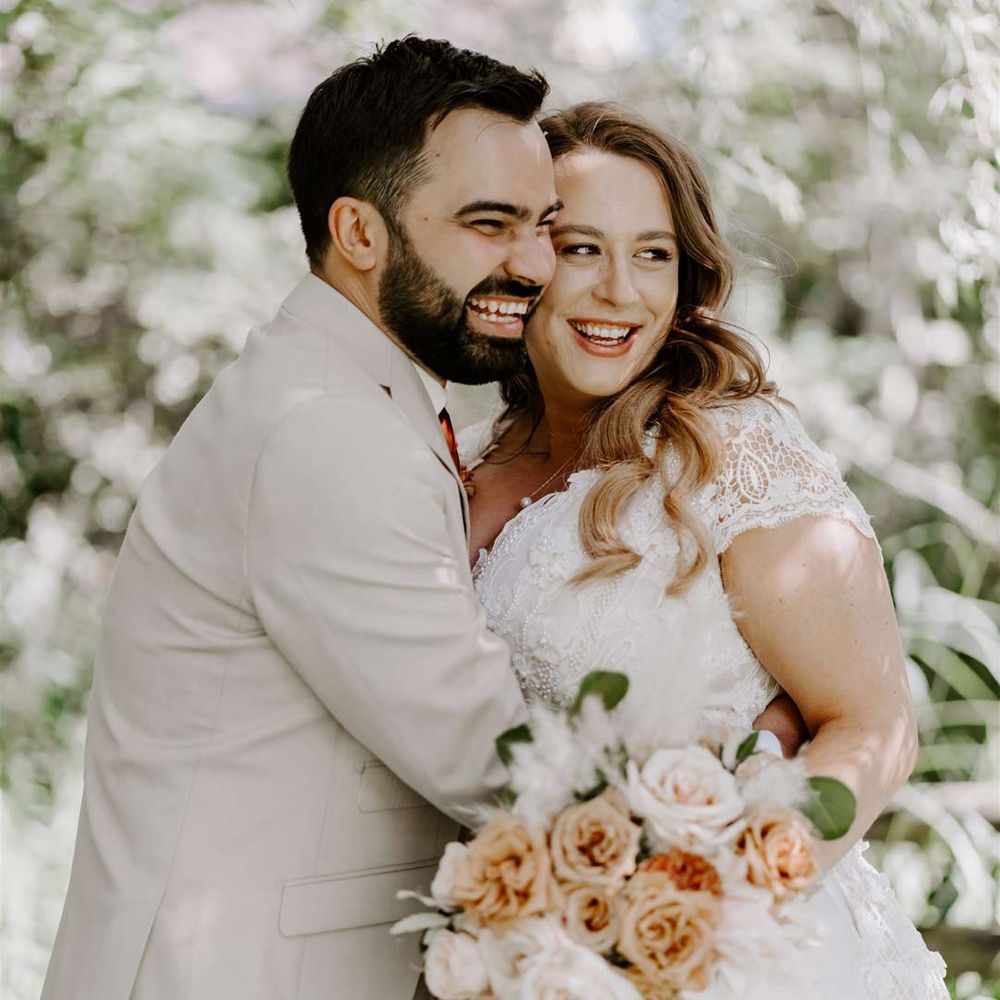 Groom in light grey suit and bride in DIY short sleeved lace wedding dress embracing at garden wedding