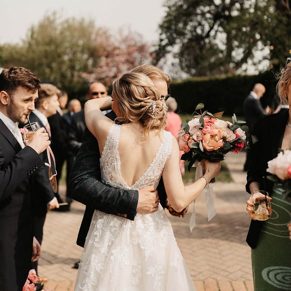 Bride in a lace wedding dress with an elegant updo holding a pink bouquet hugs a wedding guest 