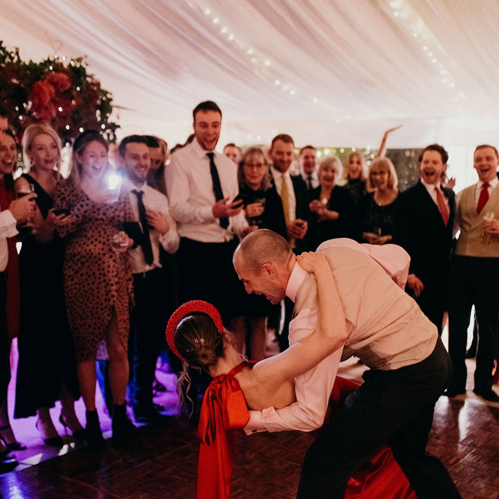 Bride in red silk wedding dress with cape, large red embellished head band, letter hair clips and large silk bow and groom in waistcoat and white shirt dancing at winter wedding party