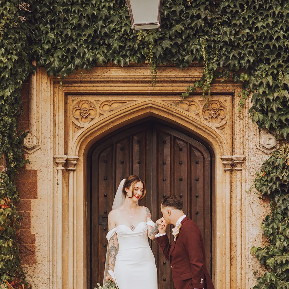 Groom in maroon suit kisses his brides hand outside St Audries Park on their wedding day