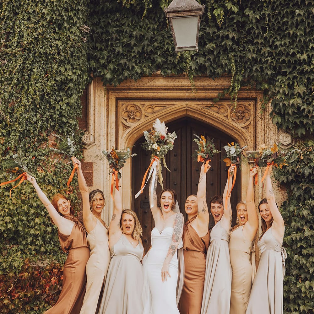 Bride lifts floral bouquet in the air with her bridesmaids in Autumnal bridesmaid dresses in mismatched styles