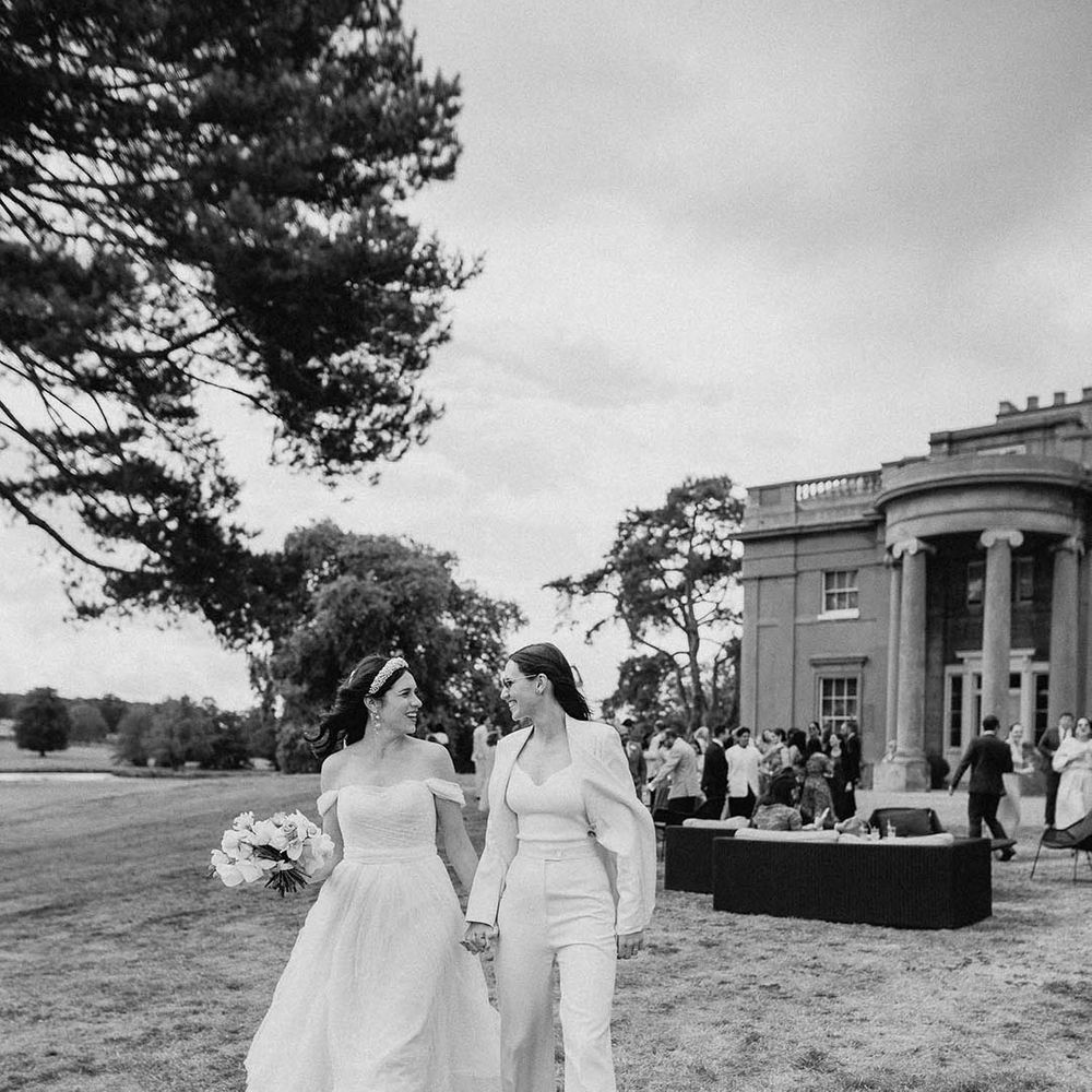 Bride in an off the shoulder wedding dress walking hand in hand with the bride in a white bridal suit 