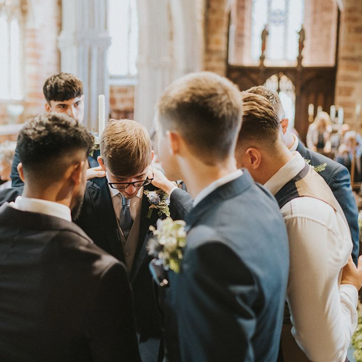 The groomsmen all pray around the groom before the wedding ceremony in matching navy suit jackets 