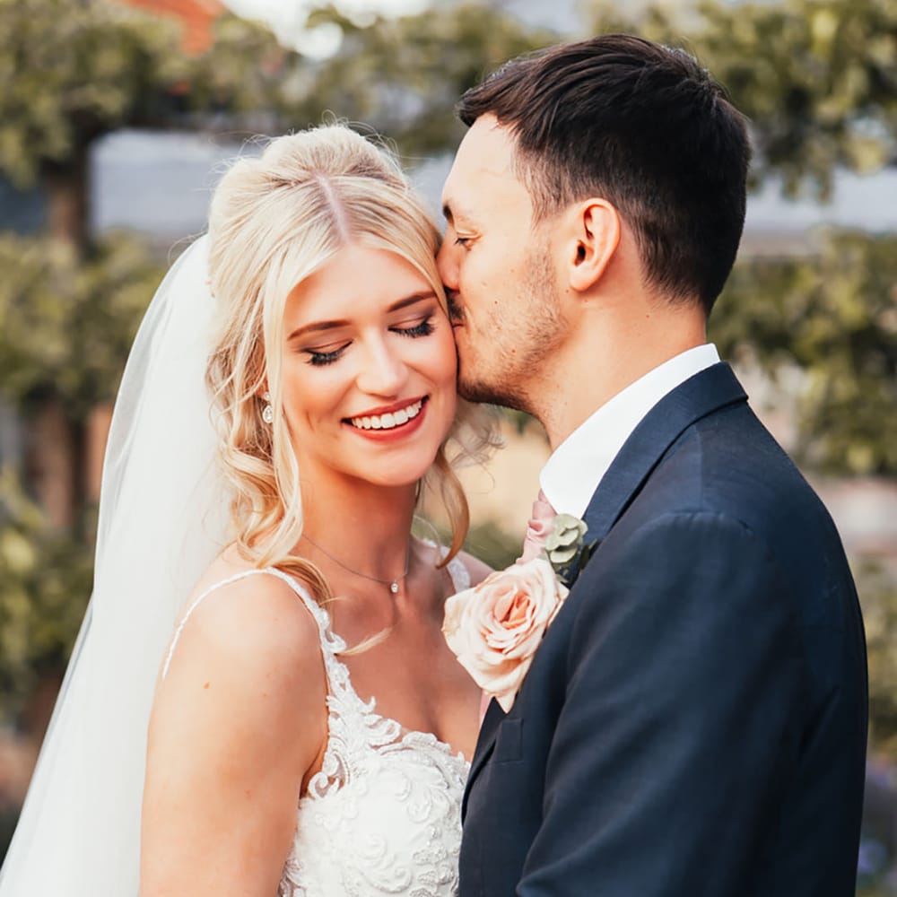 The groom in a navy suit kissing the bride on the forehead who wears a lace wedding dress and veil 