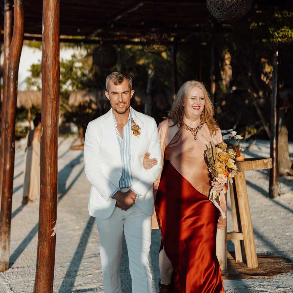Groom walking down the aisle wearing white mens wedding suit, light blue and white striped shirt, dusky orange garden rose and dried flower boutonniere, white suit trousers and gold jewellery with wedding guest in split style burgundy and dusky orange sleeveless dress 
