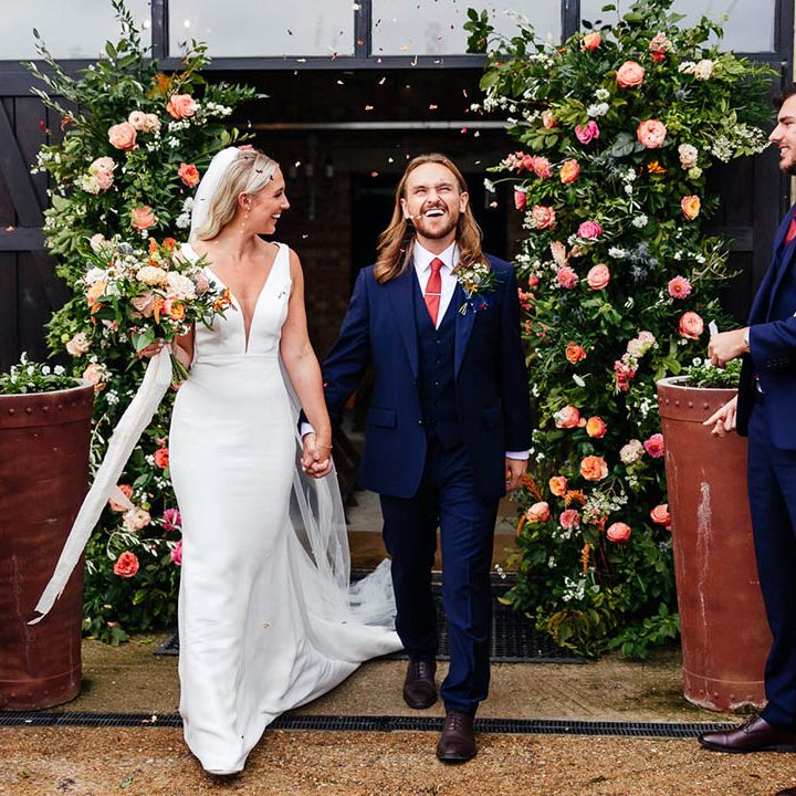 Groom in three piece blue suit walks out of the ceremony to confetti with the bride in a Savin London wedding dress 