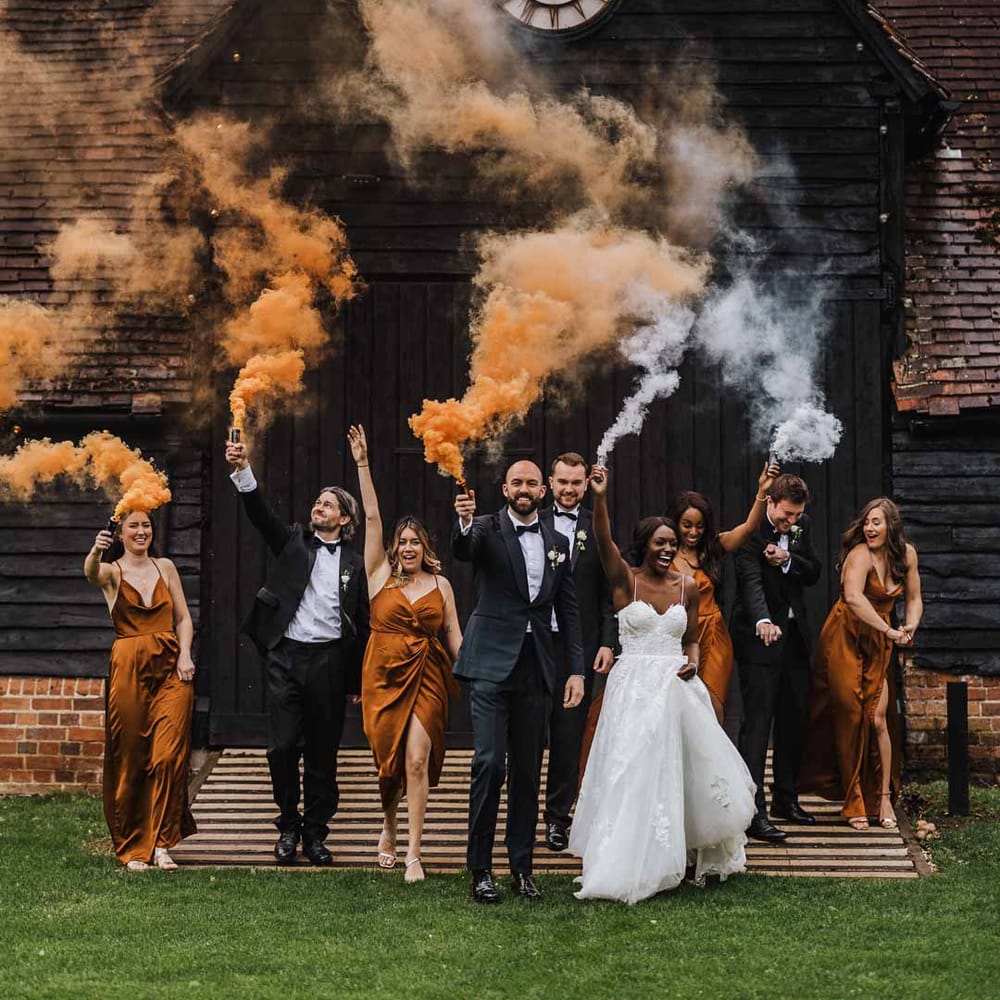 Bride in sweetheart neckline lace wedding dress with detachable off the shoulder sleeves, groom in classic black three piece grooms suit with bow tie and white rose and foliage boutonniere in church, bridesmaids wearing strappy sleeveless front slit burnt orange bridesmaid dresses holding foliage bouquets and groomsmen wearing classic black suits doing burnt orange and white smoke flares at Lains Barn wedding