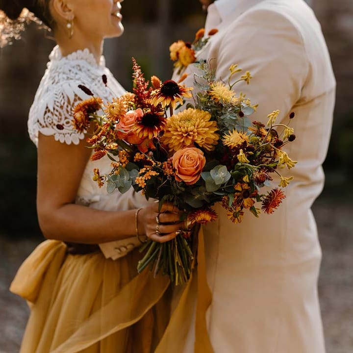 Groom in cream morning suit embracing bride in white mesh lace bridal crop top and yellow wedding skirt holding dried autumnal wedding bouquet with sunflowers, garden roses, pampas grass, eucalyptus, yellow tall kangaroo paws, orange osmanthus flowers, foliage and dried flowers at Broadfield Court wedding