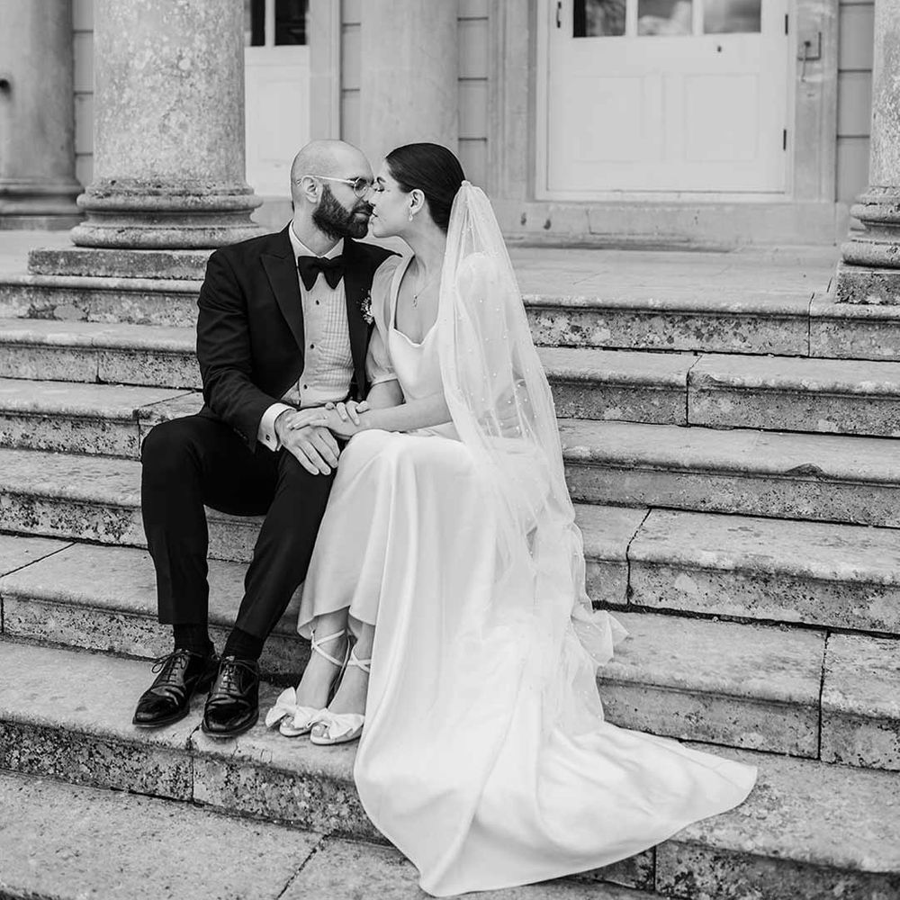 Bride in short sheer puff sleeve wedding dress with square neck and wrist loop with sheer pearl embellished bridal veil kissing groom in classic black tuxedo with bowtie and wildflower boutonniere sitting on the steps of Buxted Park