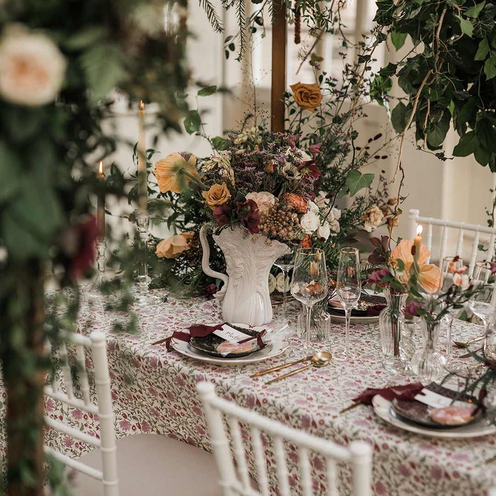 Botanical wedding tablescape with patterned floral tablecloth, twisted tapered candles, art deco vases with dried wildflower wedding centrepieces surrounded by suspended wildflower and foliage arrangements in large gold vases at Buxted Park