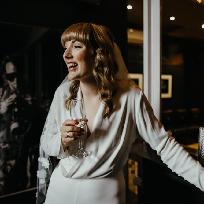 Bride in a long sleeve jumpsuit, with fringe and wavy hair enjoying a drink 