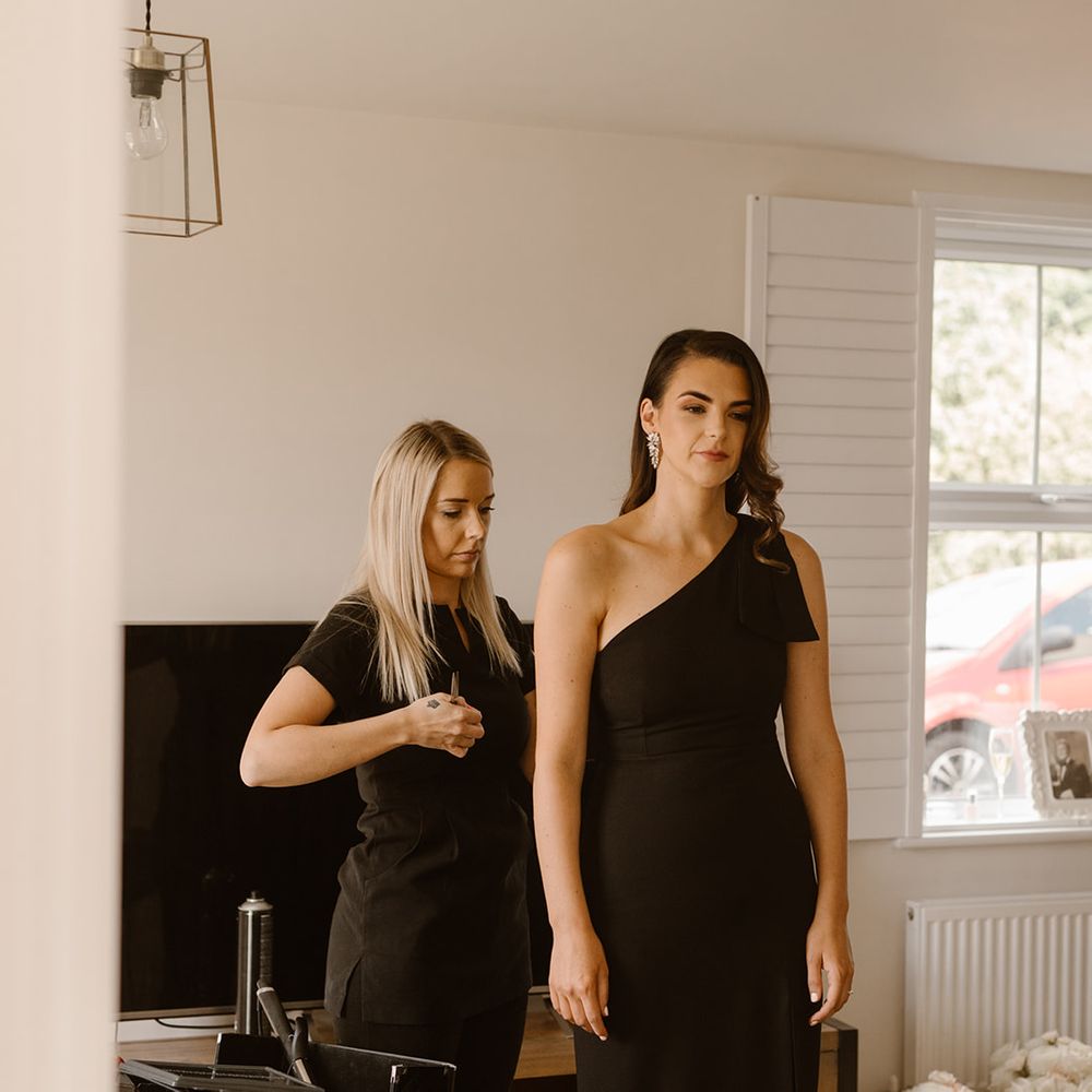 Bridesmaid in black off the shoulder bridesmaid dress with hair stylist in black tunic touching up hair before wedding