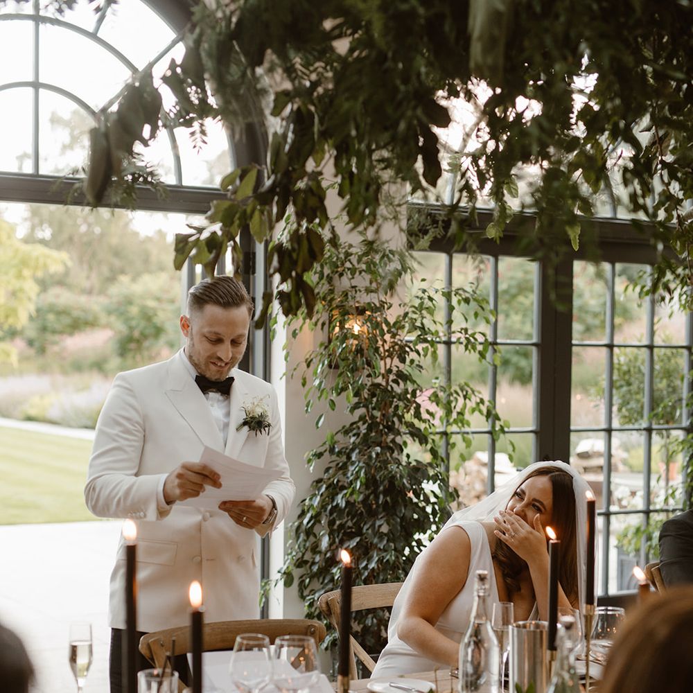 Groom in double breasted white tuxedo jacket giving speech in the Fig House at Middleton Lodge whilst bride in white Made With Love wedding dress looks up at him