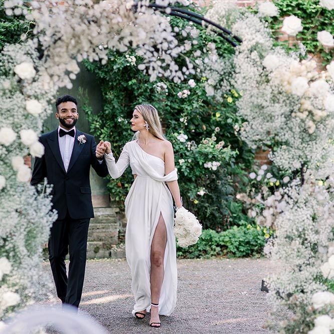 Groom in a black tuxedo and bow tie holding hands with this bride in a one shoulder wrap wedding dress in the gardens at Garthmyl Hall wedding venue 