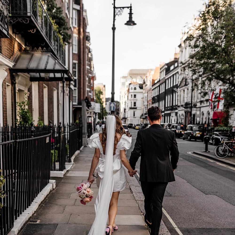 Bride in a short wedding dress with puff sleeves walking through Mayfair with her groom dresses in black with her long veil trailing behind them 