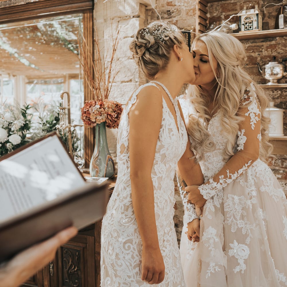 Two brides in white dresses with blonde hair kiss as a guest performs a reading at their wedding at Crab and Lobster Fairytale Wedding.