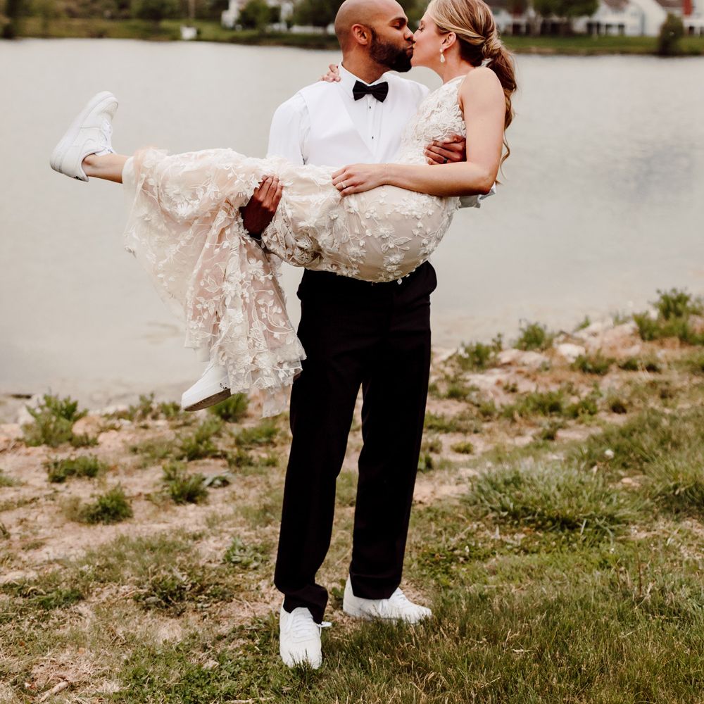 Groom in white shirt, black trousers and black bow tie carries bride in white embroidered Made With Love wedding dress as they stand next to lake both wearing white Nike Air Force trainers at backyard wedding