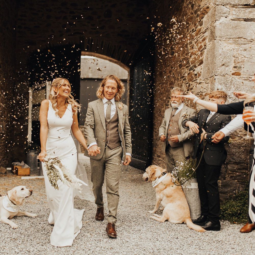 Bride in white V neck Made With Love wedding dress and veil holding white and green dried bridal bouquet walks through courtyard holding hands with groom in sage green suit and brown shoes as guests throw confetti after wedding ceremony at Anran Devon