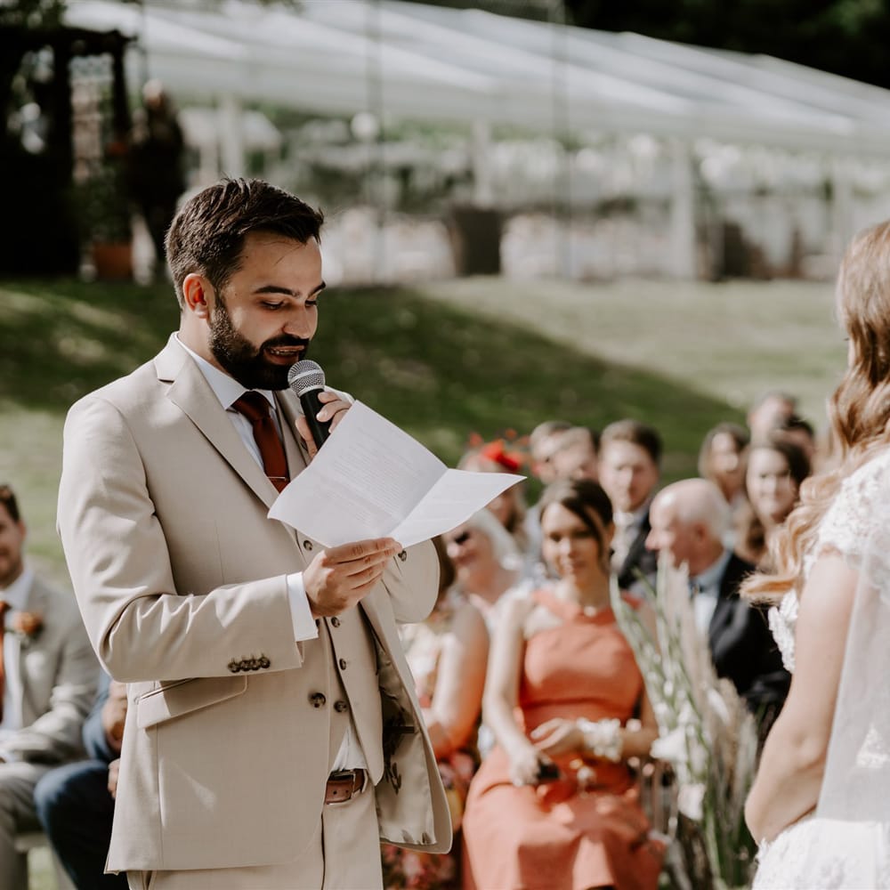 groom in beige three piece suit with burgundy tie holds microphone whilst reading out vows during DIY garden wedding ceremony in Bedfordshire