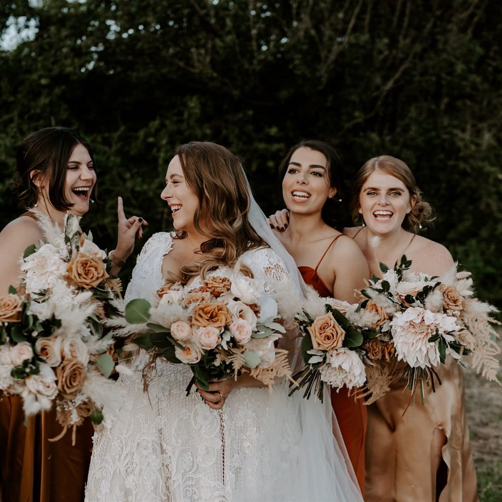 Bride in homemade lace wedding dress and veil stands with bridesmaids in assorted silk bridesmaid dresses as they all hold mixed rose wedding bouquets for outdoor wedding in Bedfordshire