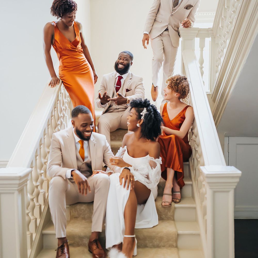 Wedding party in beige and orange suits and satin bridesmaid dresses sitting no the stairs at Modern Hall, London