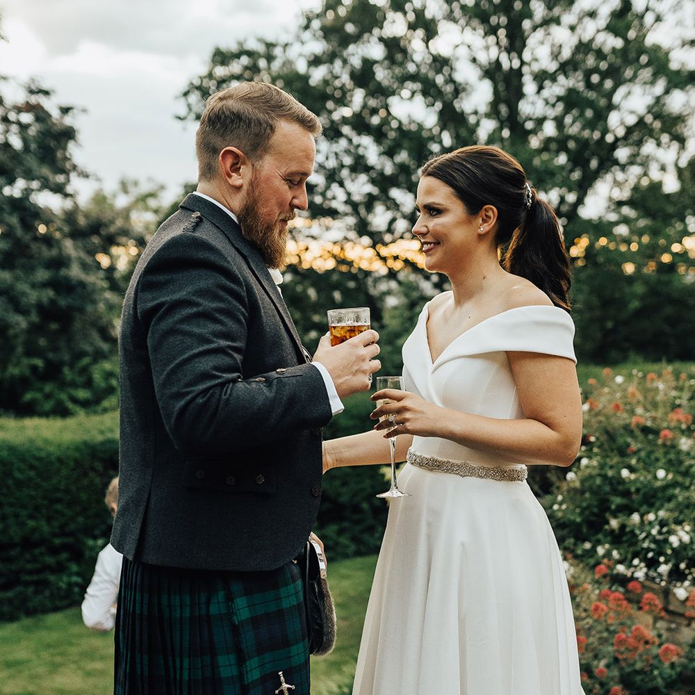 Bride in an off the shoulder Suzanne Neville wedding dress and groom in a tartan kilt drinking cocktail at sunset 