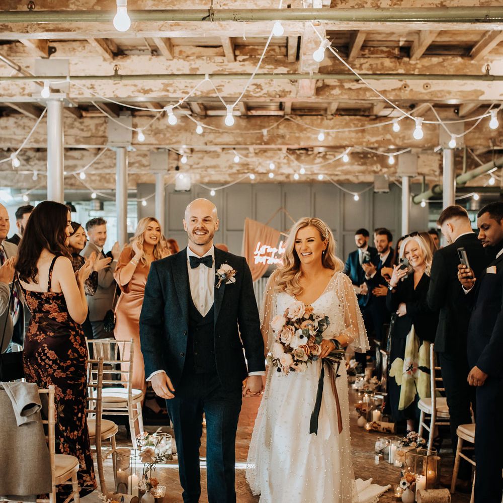 Bride in pearl wedding dress holding pastel bouquet tied with ribbon walks back down aisle with groom in three piece navy wedding suit with bow tie at industrial Mill wedding
