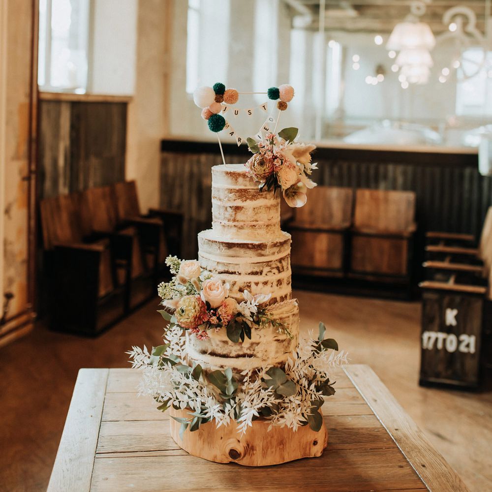 Rustic semi naked three tier wedding cake with floral decor and bunting wedding topper
