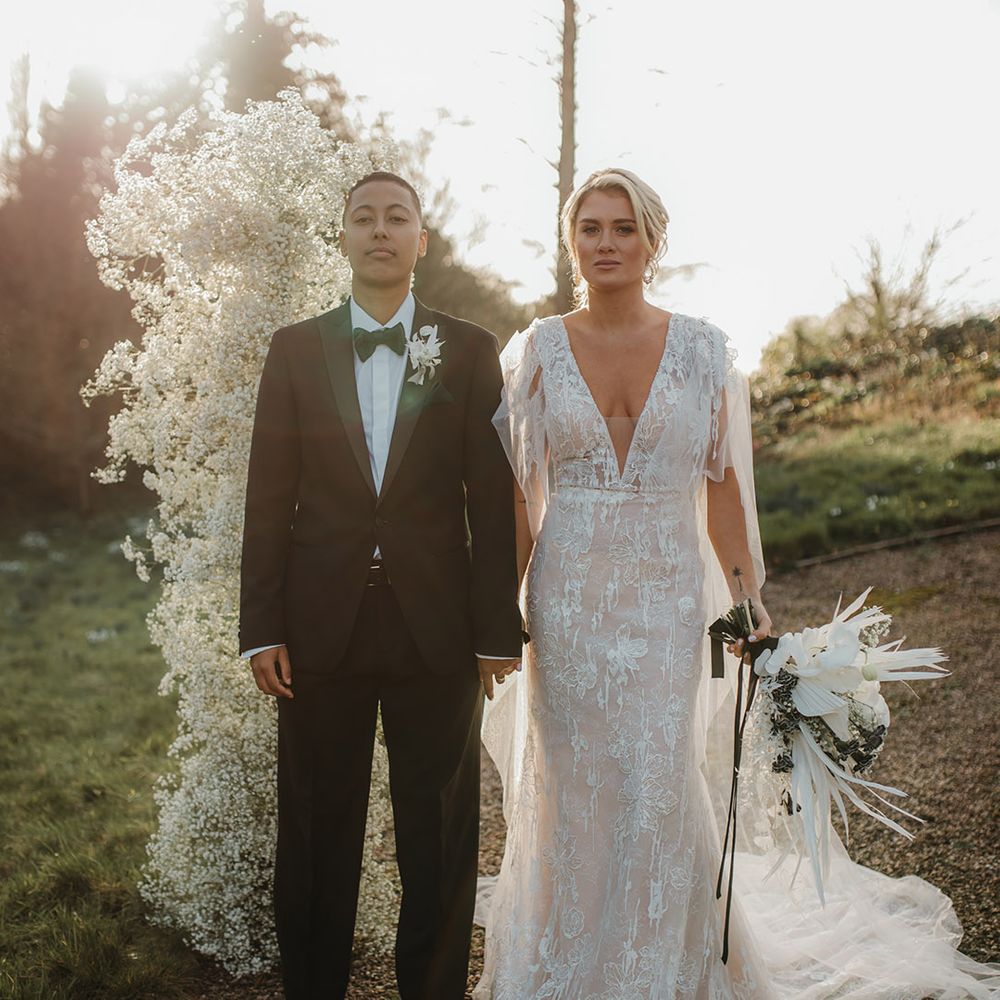 LGBTQI+ couple in a black tuxedo and lace wedding dress at golden hour with column gypsophila clouds