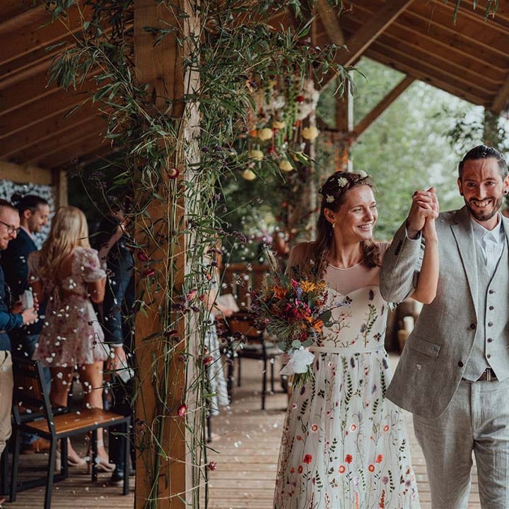 Bride and groom exit their outdoor wedding ceremony with bride wearing floral embroidered dress and groom in grey suit 