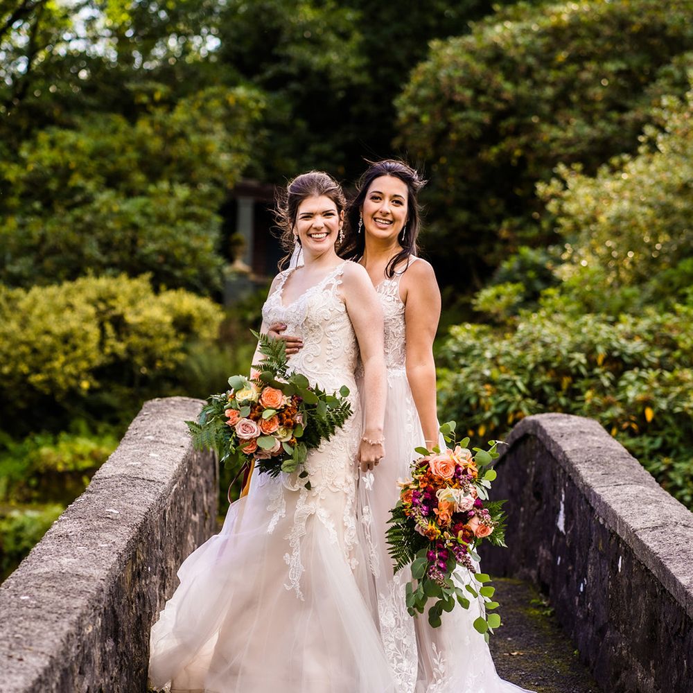 Brides stand together on bridge wearing lace wedding dresses and autumnal wedding bouquets